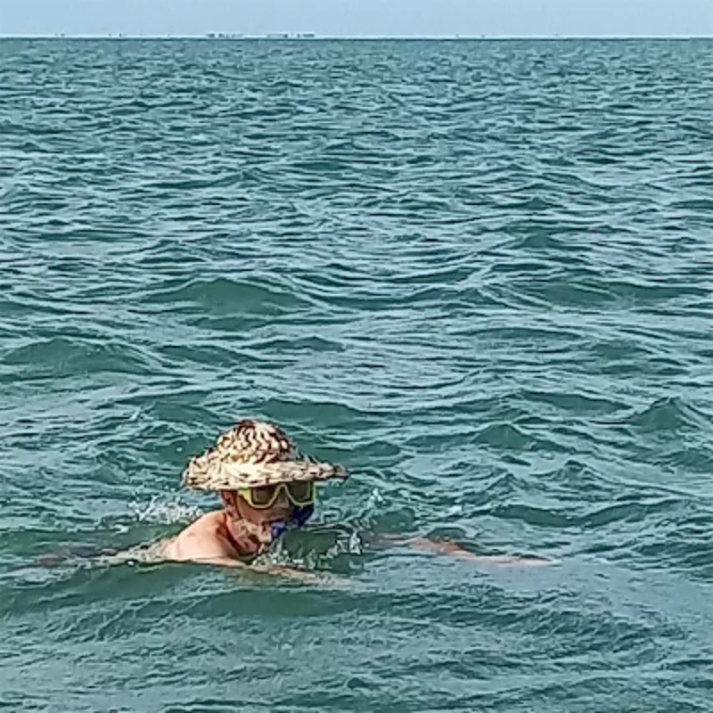 The image shows a person swimming in the ocean. The person is wearing a hat, goggles, and a snorkel. The water is choppy, and the sky is clear. In the distance, there is a faint outline of what appears to be land or structures. The overall scene suggests a day of snorkeling or swimming in the sea.